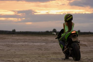 Kasklı ve yeşil Holi tozlu isimsiz bayan motosikletçi günbatımında plajda modern motosiklette oturuyor.