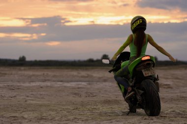 Kasklı ve yeşil Holi pudralı isimsiz bayan motosikletçi gün batımında iki parmağını deniz kıyısında gösteriyor.
