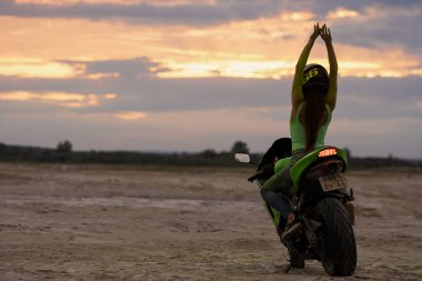 Kasklı ve yeşil Holi pudralı isimsiz bayan motosikletçi gün batımında iki parmağını deniz kıyısında gösteriyor.