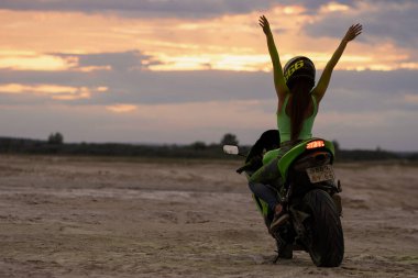 Kasklı ve yeşil Holi pudralı isimsiz bayan motosikletçi gün batımında iki parmağını deniz kıyısında gösteriyor.
