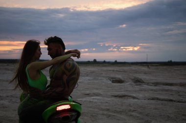 Birbirlerine aşık çiftin kumlu deniz kıyısında güneş batarken motosikletin yanında nazikçe kucaklaşmalarını yan görüyorum.