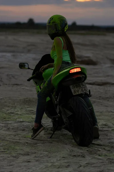 ヘルメットと緑のホリパウダーで匿名クールな女性のオートバイは日没でビーチで現代的なバイクに座っている — ストック写真
