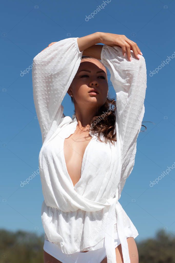 Charming young female model in stylish white blouse revealing breast standing with hands on head and looking away dreamily against blue sky in sunlight