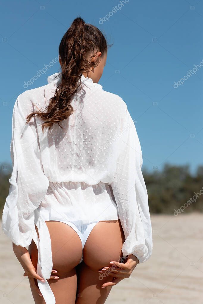 Unrecognizable female with perfect bottom pulling down white panties while standing near sandy hill on sunny day on beach