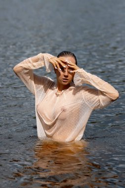 Islak bluzlu şehvetli bir kadın temiz deniz suyunda dikiliyor ve başka tarafa bakıyor.