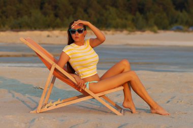 Havalı seksi esmer üstte ve bikinili iç çamaşırları gün batımında deniz kıyısında şezlongda serinlerken başka tarafa bakıyor.