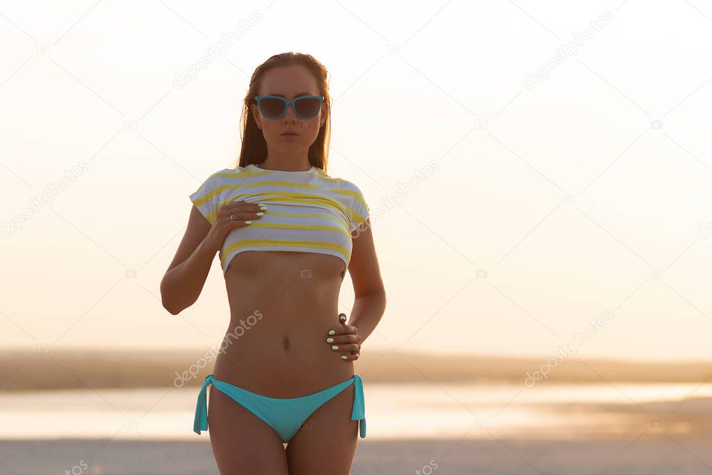 Slim young brunette wearing bikini panties with short t shirt showing sensual belly standing on beach in sunset