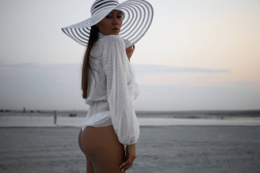 Tanga bikinili, dantelli tişörtlü, plaj şapkalı ve alacakaranlıkta kıyıda duran çekici bir kadın.