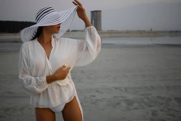 年轻性感模特身穿白色比基尼 身穿衬衫 头戴雅致的帽子 站在夕阳西下的沙滩上 — 图库照片
