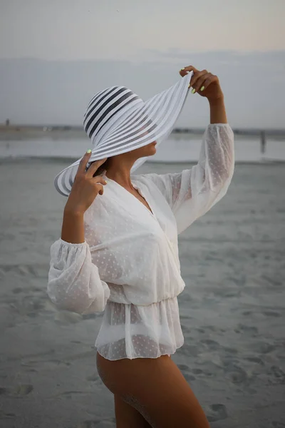 サイドビューの魅力的な女性でThongビキニとレースシャツを身に着けているビーチ帽子と立って海岸で夕暮れ — ストック写真