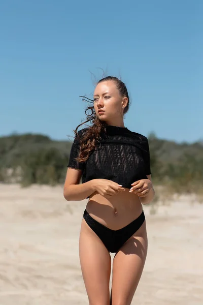 穿着时髦黑色内裤的年轻女模特的低角度 双臂交叉地站在沙滩上 与蓝天相映衬 — 图库照片