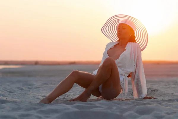 フルボディ魅力的な女性でセクシーシャツと帽子座って官能的に上の砂浜で日没光 — ストック写真