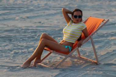 Havalı seksi esmer üstte ve bikinili iç çamaşırları gün batımında deniz kıyısında şezlongda serinlerken başka tarafa bakıyor.