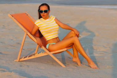 Havalı seksi esmer üstte ve bikinili iç çamaşırları gün batımında deniz kıyısında şezlongda serinlerken başka tarafa bakıyor.