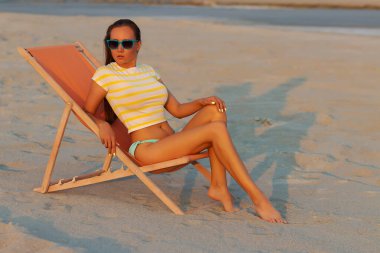 Havalı seksi esmer üstte ve bikinili iç çamaşırları gün batımında deniz kıyısında şezlongda serinlerken başka tarafa bakıyor.