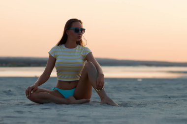 Gün batımında kumlu sahilde tek başına oturan, bikinili külotlu ve tişörtlü, ince esmer bir kız.