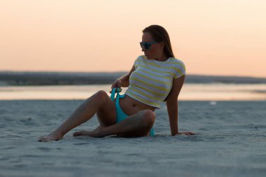 Gün batımında kumlu sahilde tek başına oturan, bikinili külotlu ve tişörtlü, ince esmer bir kız.