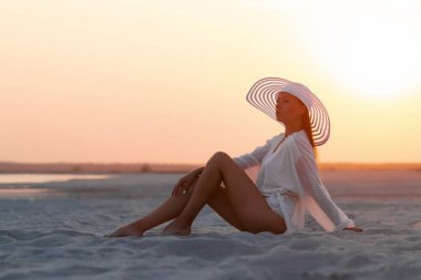 Seksi gömlekli ve şapkalı çekici kadın gün batımında kumlu sahilde şehvetle oturuyor.