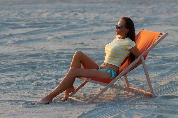 Havalı seksi esmer üstte ve bikinili iç çamaşırları gün batımında deniz kıyısında şezlongda serinlerken başka tarafa bakıyor.