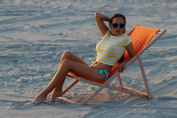 Havalı seksi esmer üstte ve bikinili iç çamaşırları gün batımında deniz kıyısında şezlongda serinlerken başka tarafa bakıyor.