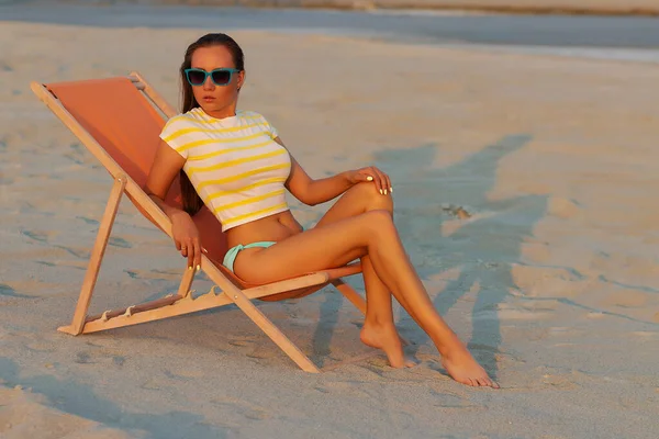 Havalı seksi esmer üstte ve bikinili iç çamaşırları gün batımında deniz kıyısında şezlongda serinlerken başka tarafa bakıyor.