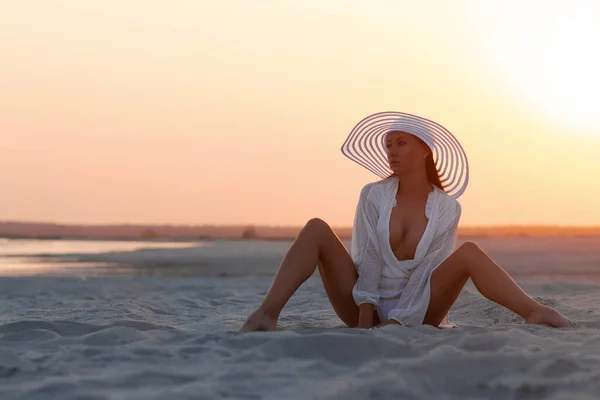 Full body alluring woman in sexy shirt and hat sitting sensually on sandy beach in sunset light