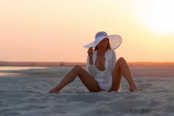 Full body alluring woman in sexy shirt and hat sitting sensually on sandy beach in sunset light