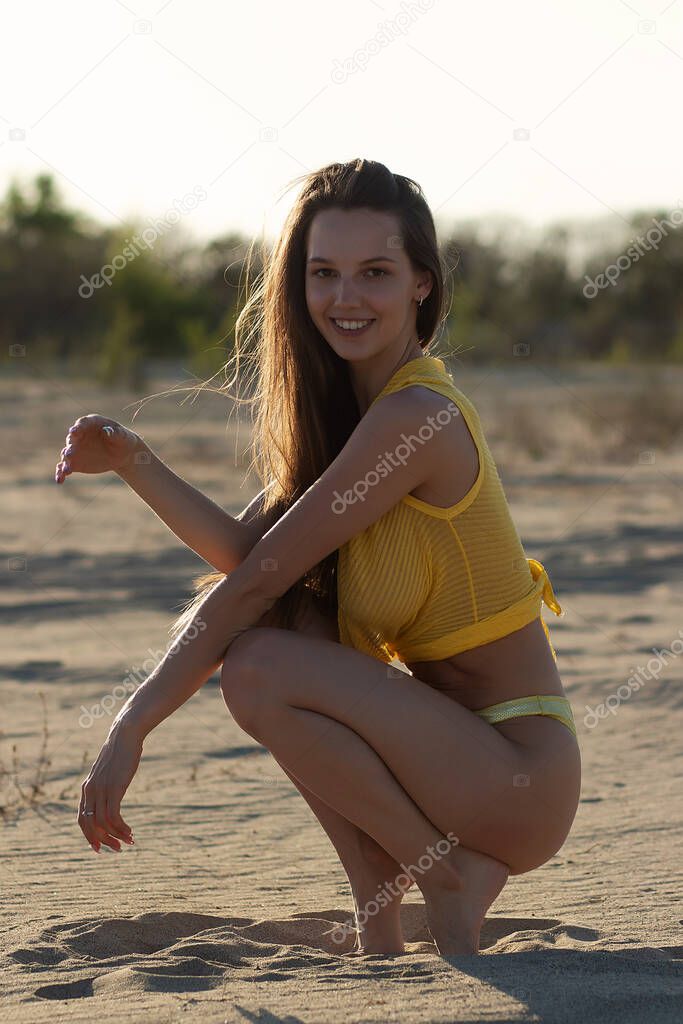 Full length of happy young female traveler with long dark hair in stylish yellow swimwear relaxing on sandy beach and looking at camera while sunbathing during summer holidays