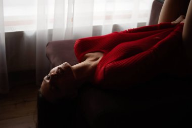 Kırmızı örgü süveter ve seksi dantel külotlu, yatakta uzanmış, şehvetli genç bir kadının yüksek açılı yan görüntüsü.