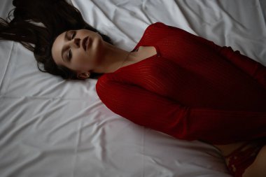 Kırmızı örgü süveter ve seksi dantel külotlu, yatakta uzanmış, şehvetli genç bir kadının yüksek açılı yan görüntüsü.
