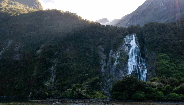 Backlit photo of waterfall surrounded by green cliffs. Location is Lady Bowen Falls located in Milford Sound, Fiordland National Park, New Zealand — Zdjęcie stockowe