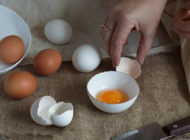 Masanın üzerinde kahverengi ve beyaz yumurtalar, bir kasede kırık bir yumurta ve dişi eller, kabuğu tekstilin üzerine koyar.