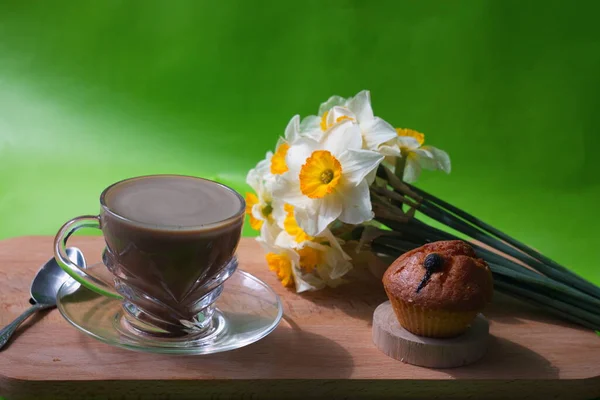 Cocoa in a transparent cup on a saucer and a cupcake that stand on a wooden table and a bouquet of white flowers