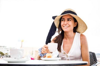 Plaj şapkalı genç Latin kadın elinde bir fincan kahve ve gülümsüyor.