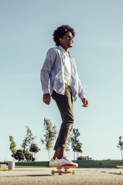 Güneş gözlüğü ve kaykay kap giyen genç koyu tenli adam resmini. Doğa arka planı.