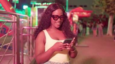 Güneş gözlüklü mutlu siyah kadın gülümsüyor ve akıllı telefonunu neon ışıklı bir lunaparkta kullanıyor.