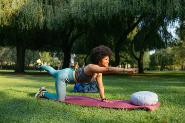 Siyah kadın dışarıda yoga yapıyor. Paspas üzerinde dengede duruyor.