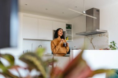 Parlak mutfağında duran bir kadın, elinde bir fincan kahveyle başka yere bakıyor.