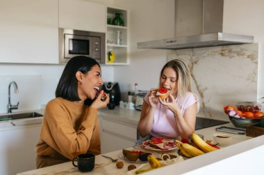 Modern bir mutfakta gülüp kahvaltı eden iki kadın.