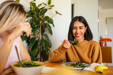 Sağlıklı bir salatanın tadını çıkaran bir kadın ve masadaki bir arkadaşıyla sohbet eden bir kadın.