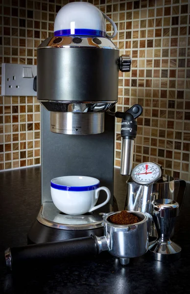 Stainless steel espresso machine with white and blue espresso cups, portafilter, ground coffee, coffee tamper, milk jug and milk thermometer in a kitchen setting with brown and tan checked tiles