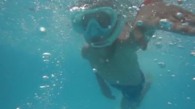 Büyük gözlüklü küçük bir çocuk denizin altında kameraya doğru yüzüyor.