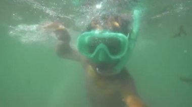 Büyük gözlüklü küçük bir çocuk denizin altında kameraya doğru yüzüyor.