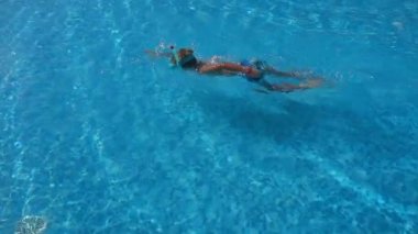 Çocuk havuzda yüzer, maske takar, koyu mavi renkte.