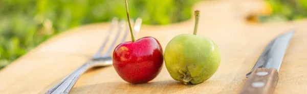 An apple and a cherry are lying on a cutting board, next to a knife and fork on a background of green grass. The concept of a diet, eco-food, separate nutrition, veganism, a healthy lifestyle. — Stock Photo, Image