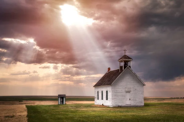 Old rural church in the countryside at sunset with sun rays shining down from the clouds. There is a small outhouse in the picture. This looks like a church from the old western days.