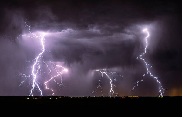 A fierce lightning storm with lots of cloud to ground lightning strikes shooting from the clouds. There clouds are purple with stream of rain falling downward towards the ground.