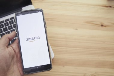 Madrid İspanya, 13 Haziran 2021; Eller ekranında Amazon uygulamasının logosu ve arka planında bilgisayar olan akıllı bir mobil cihaz tutuyor..