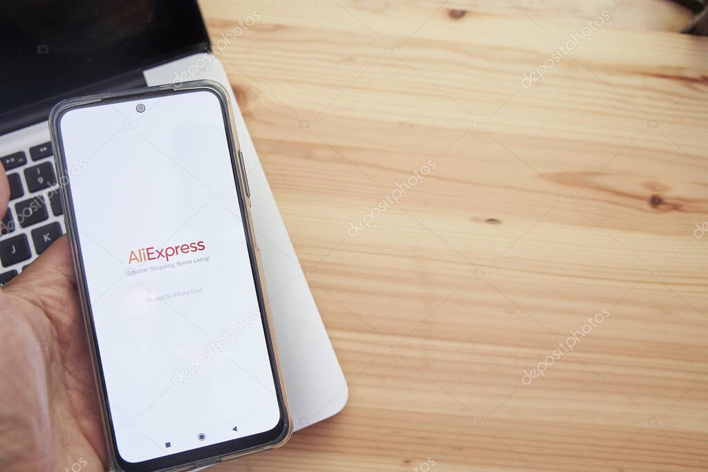 Madrid Spain, June 13 2021; Hands holding a smart mobile device with the logo of the company's AliExpress application on the screen and a computer in the background.