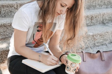 Bir fincan kahveye not defterine yazan bir kadın, merdivenlerde oturuyor. Yaşam tarzı kavramı